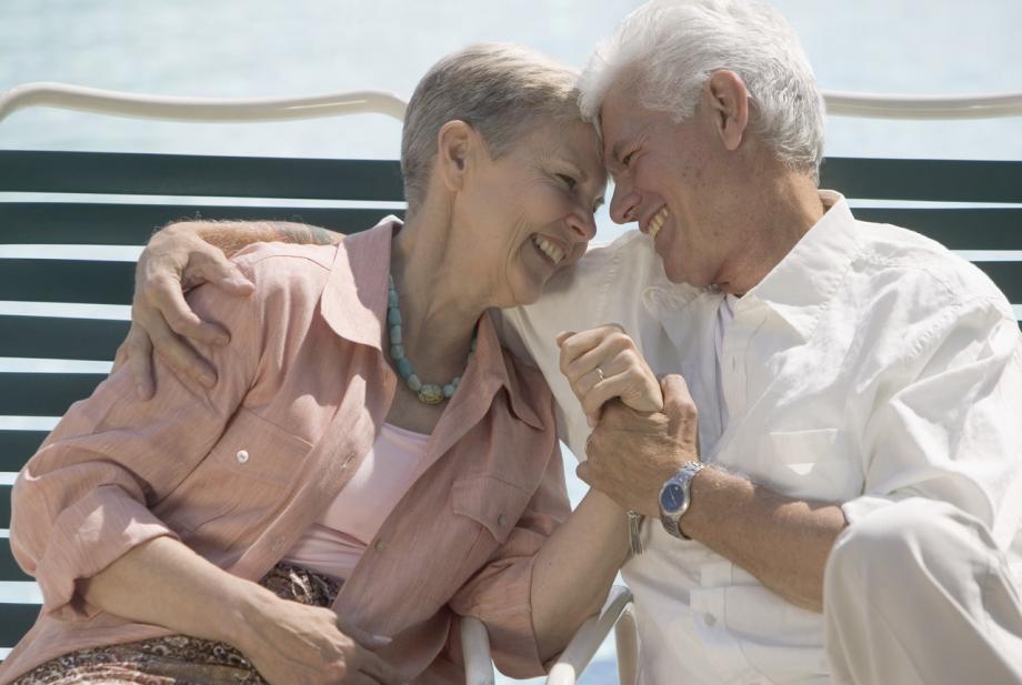 To pensjonister som sitter på en benk, smiler til hverandre og holder hverandre i hånda.