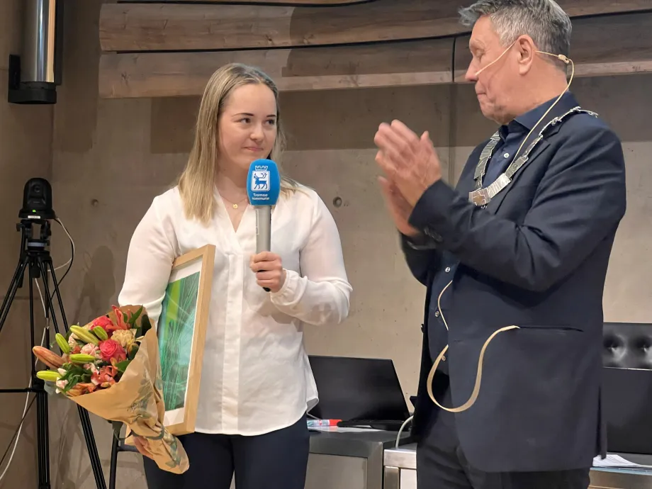 Vilde Nilsen st&aring;r med blomsterbukett og kunstverk i h&aring;nda. Holder tale via mikrofon. Ordf&oslash;reren st&aring;r ved siden av og applauderer.