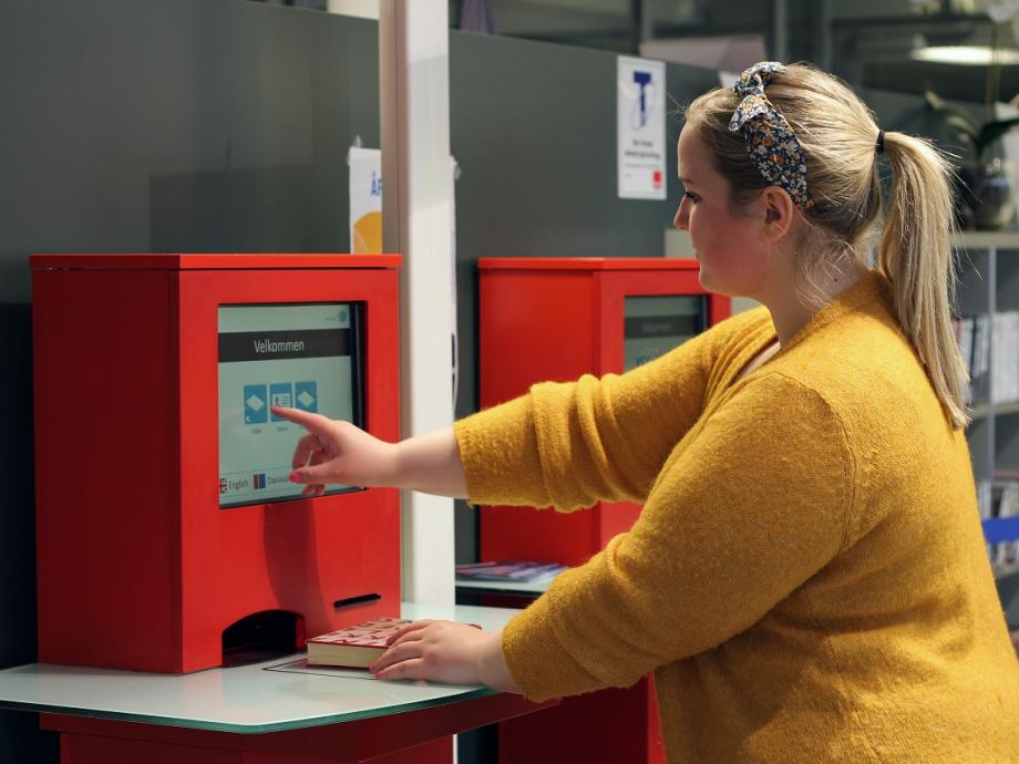 Kvinne i gul genser trykker på skjermen på en rød utlånsautomat. Foto.