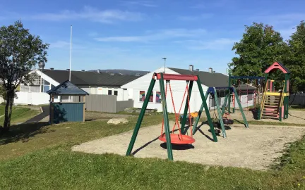 Uteomr&aring;de i Kroken barnehage med husker, klatret&aring;rn, sandkasse, gr&oslash;nt gress og tr&aelig;r en sommerdag med bl&aring; himmel 
