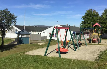 Uteområde i Kroken barnehage med husker, klatretårn, sandkasse, grønt gress og trær en sommerdag med blå himmel 