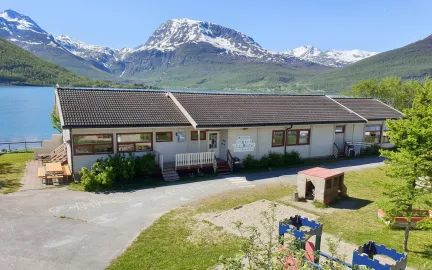 Lakselvbukt barnehage sett utenfra med lekeomr&aring;de, hav og fjell en str&aring;lende sommerdag