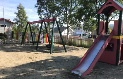 Lekeområdet i Slettaelva barnehage med høye trær, husker og klatretårn en solrik sommerdag