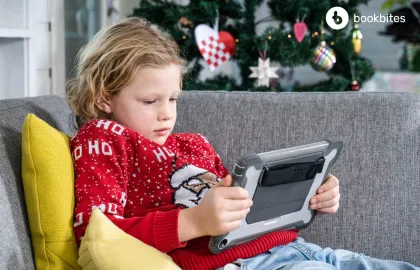 Barn i rød genser med nisse på ser på et nettbrett. I bakgrunnen står et juletre. Foto.