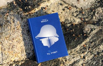Ei blå bok med en hvit illustrasjon av et menneskehode med hatt. Boka ligger på et berg. Tittelen er "Til fyret". Foto.