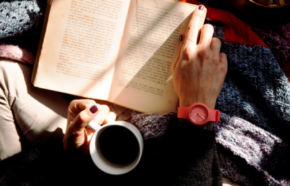 Utsnitt sett ovenfra av noen som blar i en bok med høyre hånd og holder en kaffekopp i venstre. Foto.