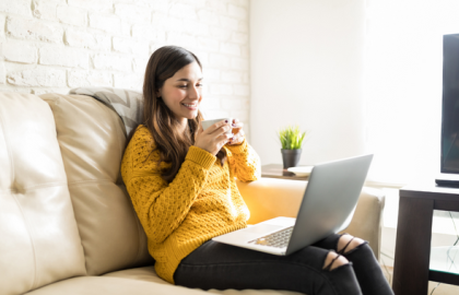 Kvinne i gul genser holder en kopp og ser på en PC hun har i fanget. Foto.