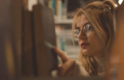 Kvinne med blondt hår og runde briller ser på bøker i en hylle. Foto.