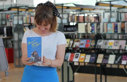 Kvinne i hvit t-skjorte og blått skjørt smiler og ser ned mot ei blå bok hun holder foran seg. I bakgrunnen er det bokhyller. Foto.