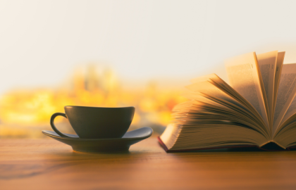 Svart kopp på svart tefat står ved siden av en oppslått bok. Foto.