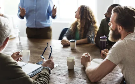 En gruppe mennesker sitter rundt et m&oslash;tebord og lytter til en person som st&aring;r og snakker.