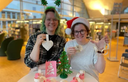 To bibliotekarer smiler mot kamera mens de holder opp julepynt laget av boksider. En har grønt år og ei hårbøyle med juletrær på. Den andre har brunt hår og nisselue på. Foto