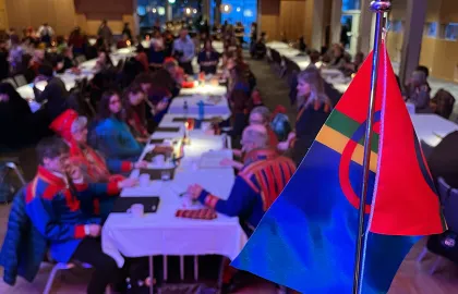 Samisk flagg på talerstolen i Tromsøsalen under samenes nasjonaldag 2024 på Radisson hotell. Koftekledde folk sitter i bakgrunnen.