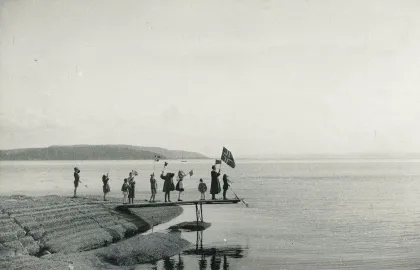 Fra frigjøringsdagene, mai 1945 Foto: Folke Beckman Mennesker står ute på en kai/et stupebrett og vifter med det norske flagget. I bakgrunnen sees hav og en lav fjellformasjon. Svart-hvitt-forto.