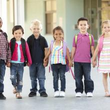 Barneskolebarn står oppstilt i skolekorridoren med ryggsekker på. Smiler til fotografen.