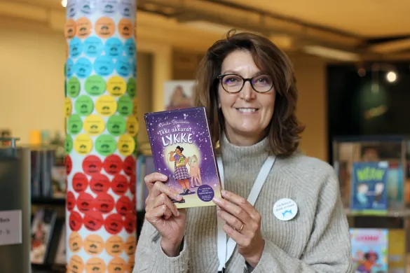 En bibliotekar smiler og holder opp boka "Ikke akkurat lykke"