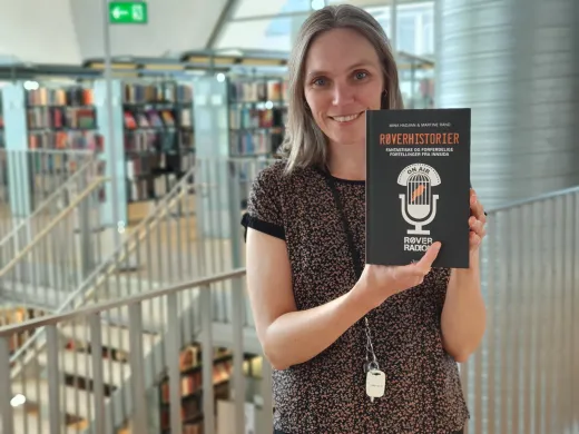 Dame med skulderlangt h&aring;r og m&oslash;nstrete bluse holder boka R&oslash;verhistorier og smiler mot kamera. Foto.