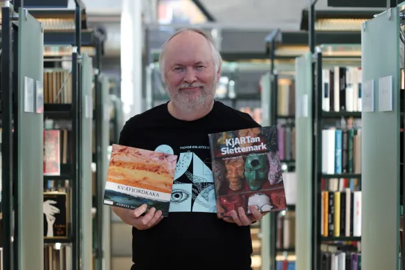 Mann i svart skjorte med lysebl&aring;tt trykk holder opp to b&oslash;ker og smiler mot kamera. Foto.