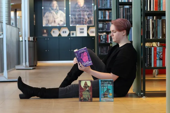 Kvinne sitter p&aring; gulvet og leser ei bok med ryggen lent mot ei bokhylle. Hun er kledd i svart og har lange, svarte st&oslash;vletter p&aring; seg. Foran henne st&aring;r to b&oslash;ker med fronten mot kamera. Foto.