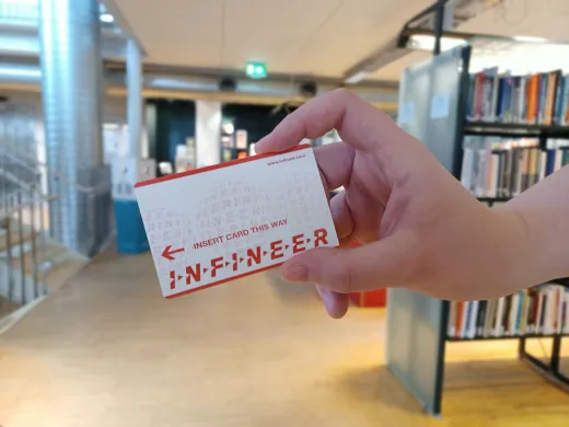 Et hvitt og oransje kort holdes av en h&aring;nd. I bakgrunnen st&aring;r det bokhyller. Foto.