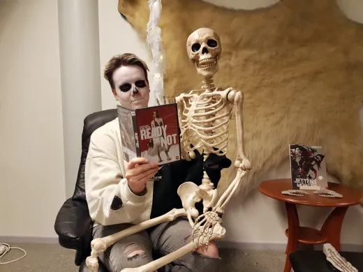 Kvinne med Halloweensminke i ansiktet sitter med et leke-skjelett p&aring; fanget. Hun holder opp et filmdeksel. Foto