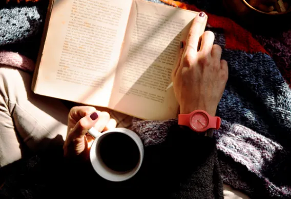 Utsnitt sett ovenfra av noen som blar i en bok med h&oslash;yre h&aring;nd og holder en kaffekopp i venstre. Foto.