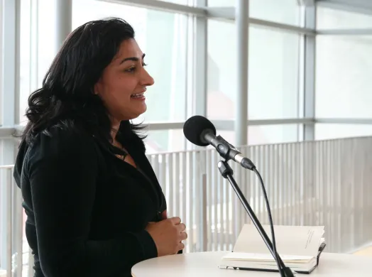 Kvinne med svart h&aring;r og svarte kl&aelig;r snakker i en mikrofon. Foto