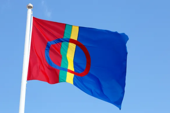 Det samiske flagget p&aring; flaggstang mot bl&aring; himmel. Foto.