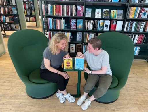 To kvinner ler mot hverandre og har to b&oslash;ker st&aring;ende mellom seg p&aring; et lite bord. De sitter i gr&oslash;nne lenestoler med bokhyller bak seg. Foto.