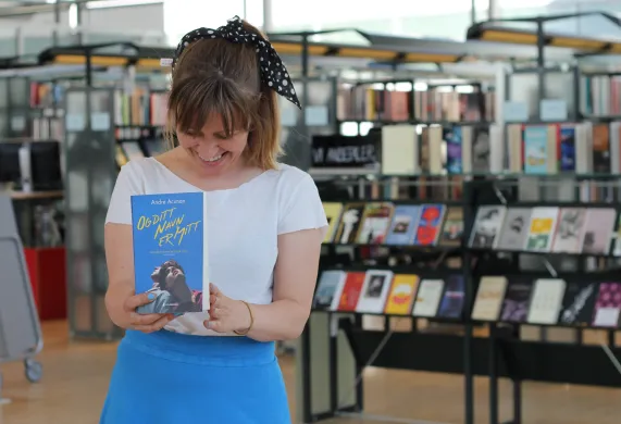 Kvinne i hvit t-skjorte og bl&aring;tt skj&oslash;rt smiler og ser ned mot ei bl&aring; bok hun holder foran seg. I bakgrunnen er det bokhyller. Foto.