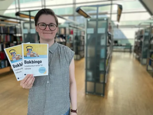 Kvinne med kort brunt h&aring;r, briller og gr&aring;-og-hvit-stripete skjorte holder to ark og smiler mot kamera. I bakgrunnen er vinduer og bokhyller. Foto.