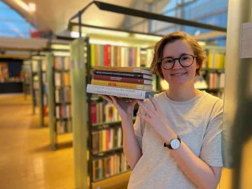Kvinne med halvlangt, brunt h&aring;r og lys gr&aring; t-skjorte holder en bunke b&oslash;ker og smiler mot kamera. I bakgrunnen st&aring;r reoler med b&oslash;ker. Foto