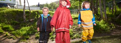 Tre barn med regntøy i Fjellvegen barnehage en sommerdag med grønt gress og skog